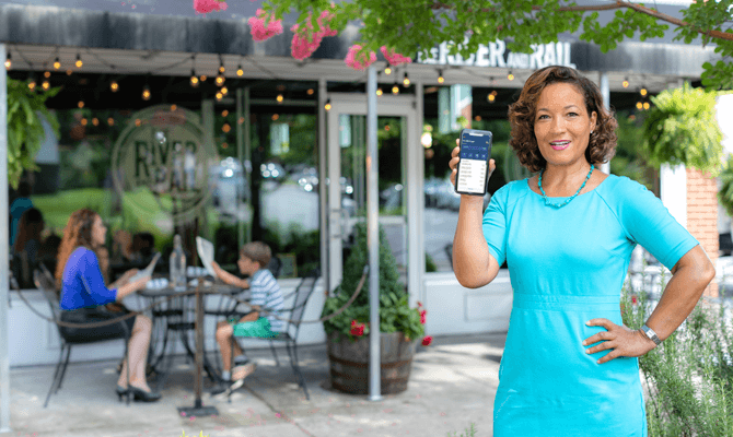 Woman holding smartphone up with mobile banking screen showing in front of River and Rail restaurant