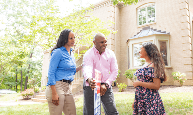 Smiling family of three looking at a sold sign in front of a house