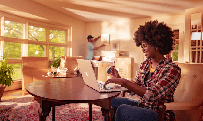 Smiling woman holding credit card and using laptop while man hangs artwork in the background