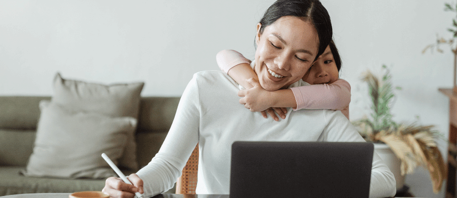 Mother working on laptop while her young daughter hugs her from behind