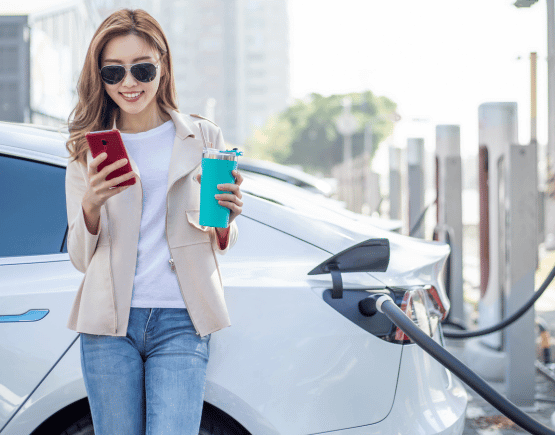 Young smiling woman holding tumbler and using smartphone while charging her car